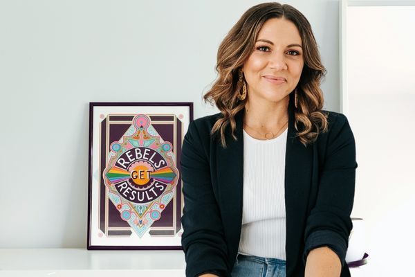 Verity White, founder of Checklist Legal with light brown hair standing in front of an art print that reads "Rebels Get Results"